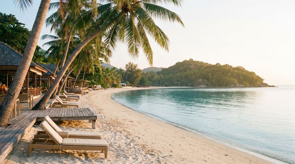 A calm Koh Samui beach-town bay at golden hour, pale sand, palms, empty wooden loungers and a distant green headland