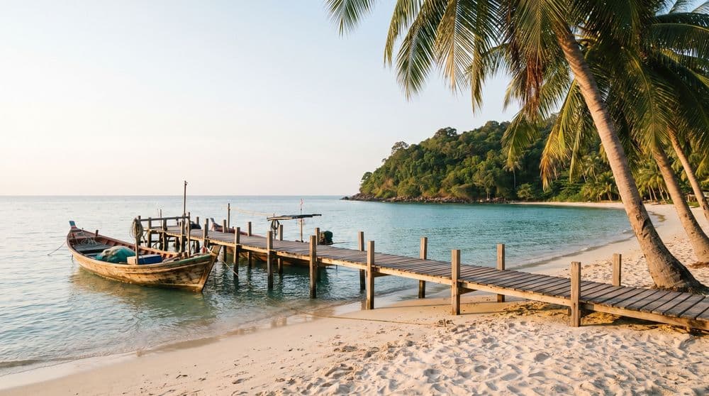Golden-hour wooden jetty over calm turquoise water toward a distant green headland, small fishing boats moored alongside, Koh Samui