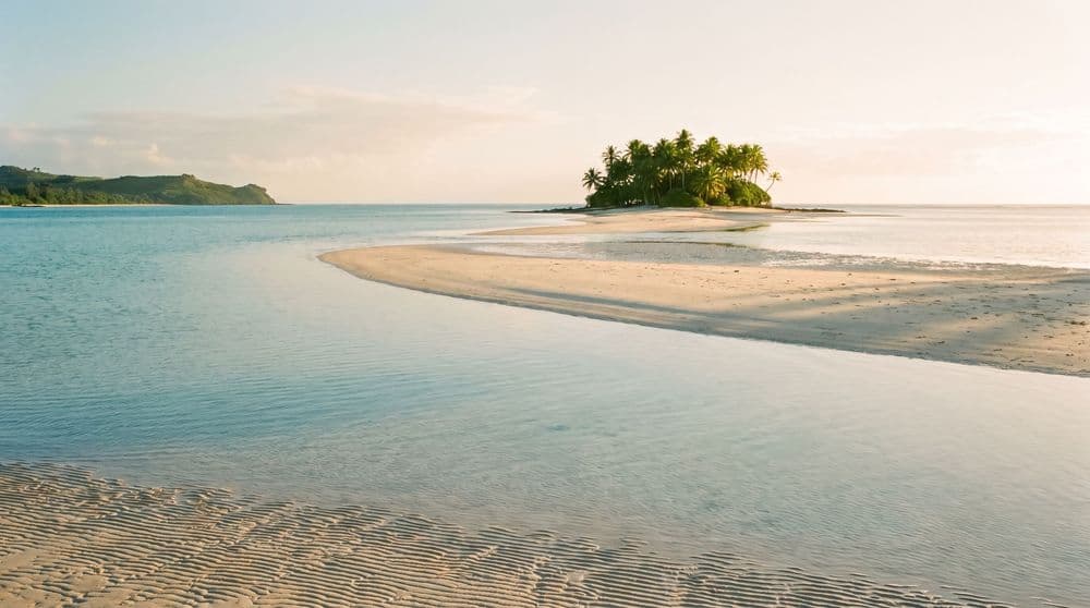 A small palm-covered islet just offshore in calm shallow turquoise water at golden hour, with a pale sandbar curving toward it