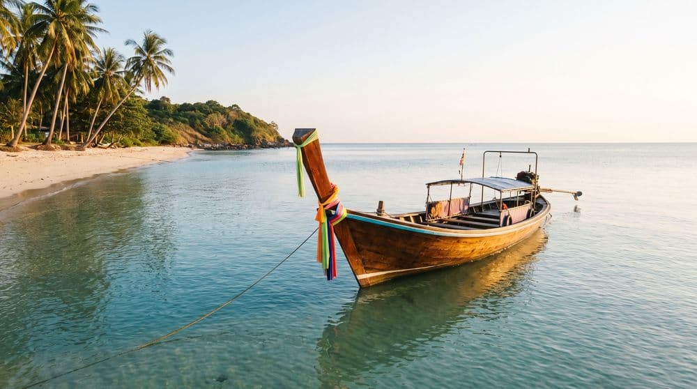 A wooden Thai longtail boat moored in calm turquoise shallows off a quiet palm-lined beach at golden hour