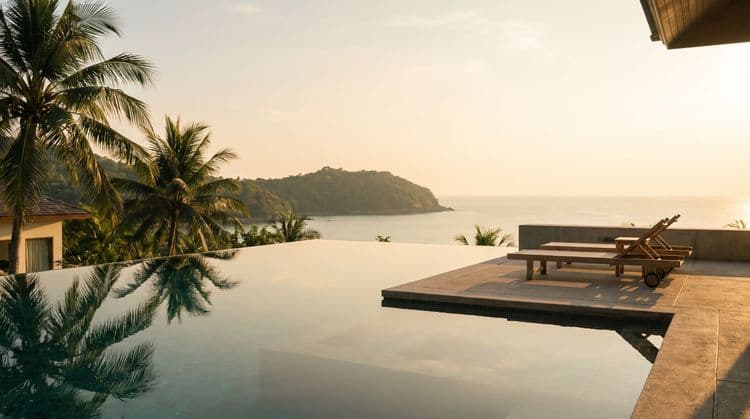 Still infinity pool on a private villa terrace at golden hour, overlooking a calm tropical bay and green headland