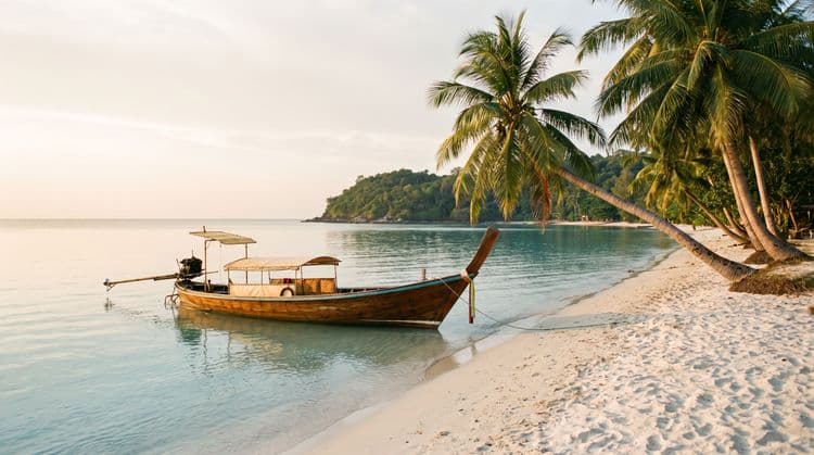 A single wooden longtail boat moored in calm golden-hour shallows off a quiet palm-lined beach