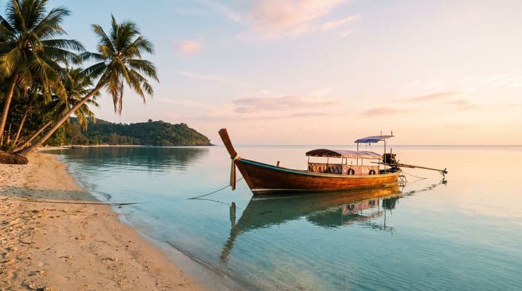 A traditional wooden longtail boat moored in calm turquoise shallows off a quiet Koh Samui beach at golden hour, palms leaning in and a green headland beyond
