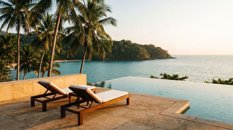 Golden-hour infinity pool edge on a private Koh Samui terrace overlooking a calm turquoise bay and green headland, two empty wooden loungers, coconut palms framing the view