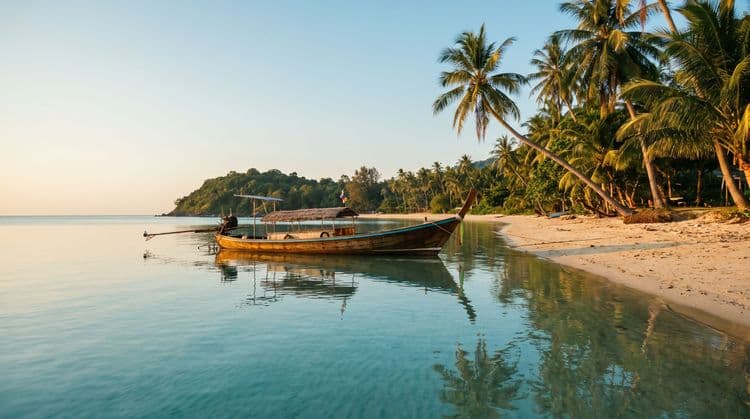 A traditional wooden longtail boat moored in calm golden-hour shallows off a quiet Koh Samui beach, seen from the water looking back toward palm-lined sand