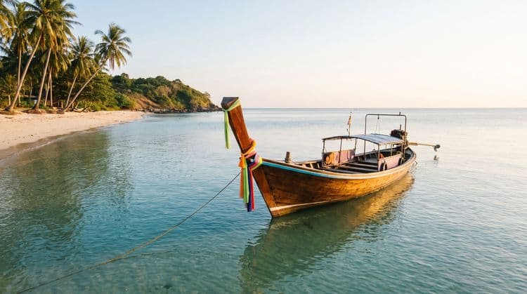A wooden Thai longtail boat moored in calm turquoise shallows off a quiet palm-lined beach at golden hour