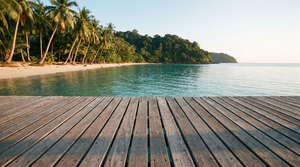 Weathered wooden deck at the edge of a calm turquoise bay at golden hour, coconut palms and a jungle headland across the water