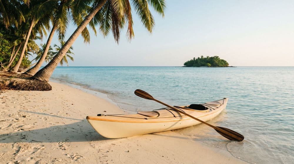 A single kayak with a wooden paddle resting on pale sand at the edge of a calm turquoise bay at golden hour, palms leaning overhead and a small green islet offshore