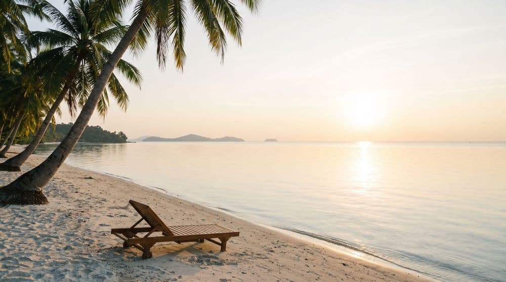 A quiet tropical bay at golden hour with a single wooden sun lounger on the sand and distant island silhouettes on the horizon