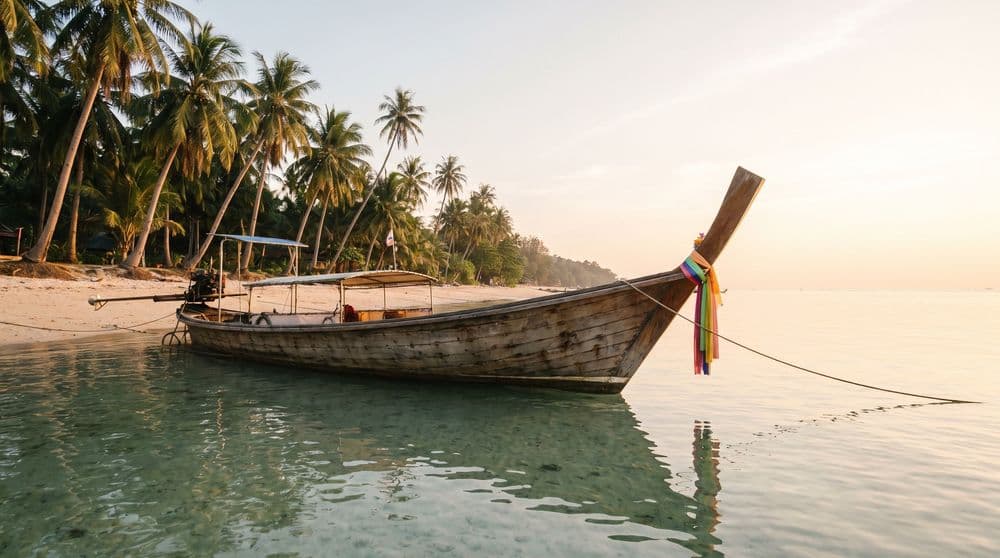 A traditional wooden longtail boat moored in calm golden-hour shallows off a palm-lined Koh Samui shore
