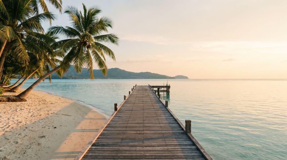 Golden-hour wooden jetty leading out over calm turquoise water toward a distant green island on the horizon, coconut palms leaning in from the shore
