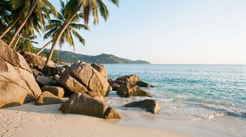 Golden-hour granite boulders meeting pale sand at the shoreline, calm turquoise surf, coconut palms and a distant green headland, Koh Samui